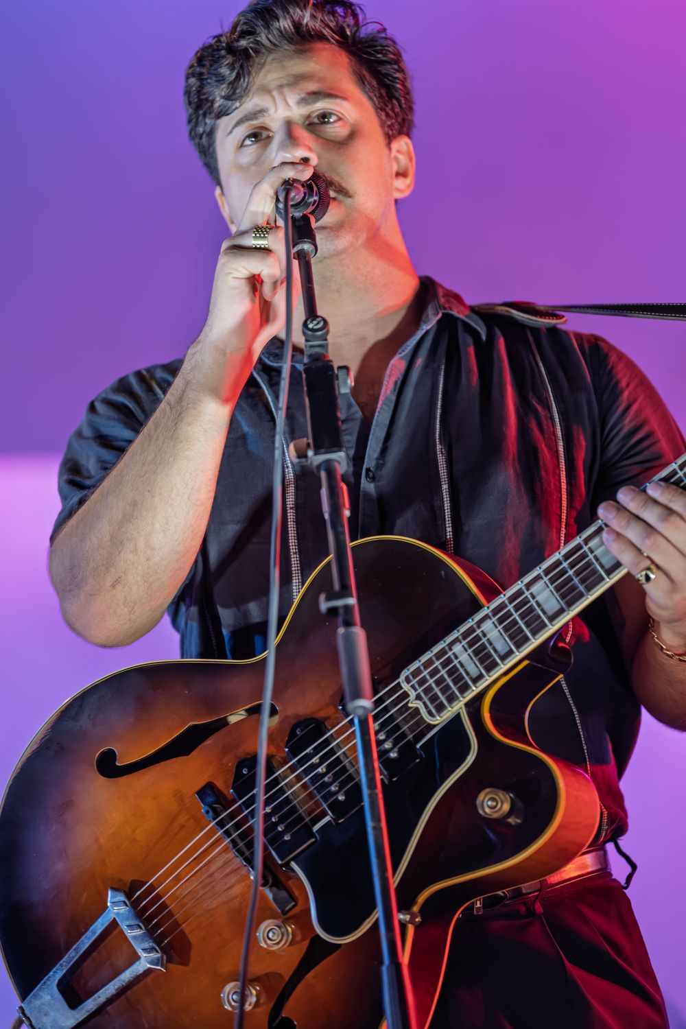 Male guitarist in black shirt performing live on stage with hollow-body electric guitar under purple and red stage lighting.
