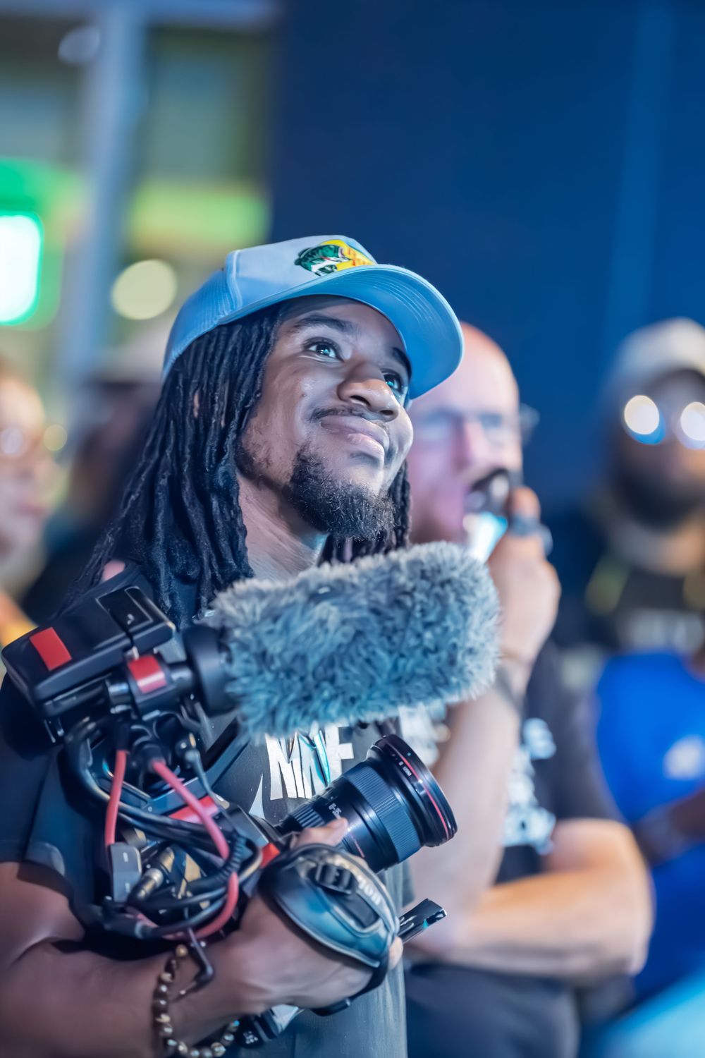 Smiling videographer with dreadlocks and blue cap holding professional camera gimbal stabilizer at an indoor event.