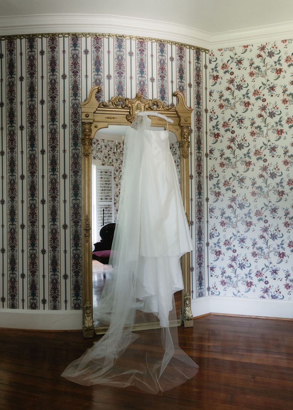 White wedding dress with veil draped over ornate gold mirror in room with floral wallpaper and hardwood floors.