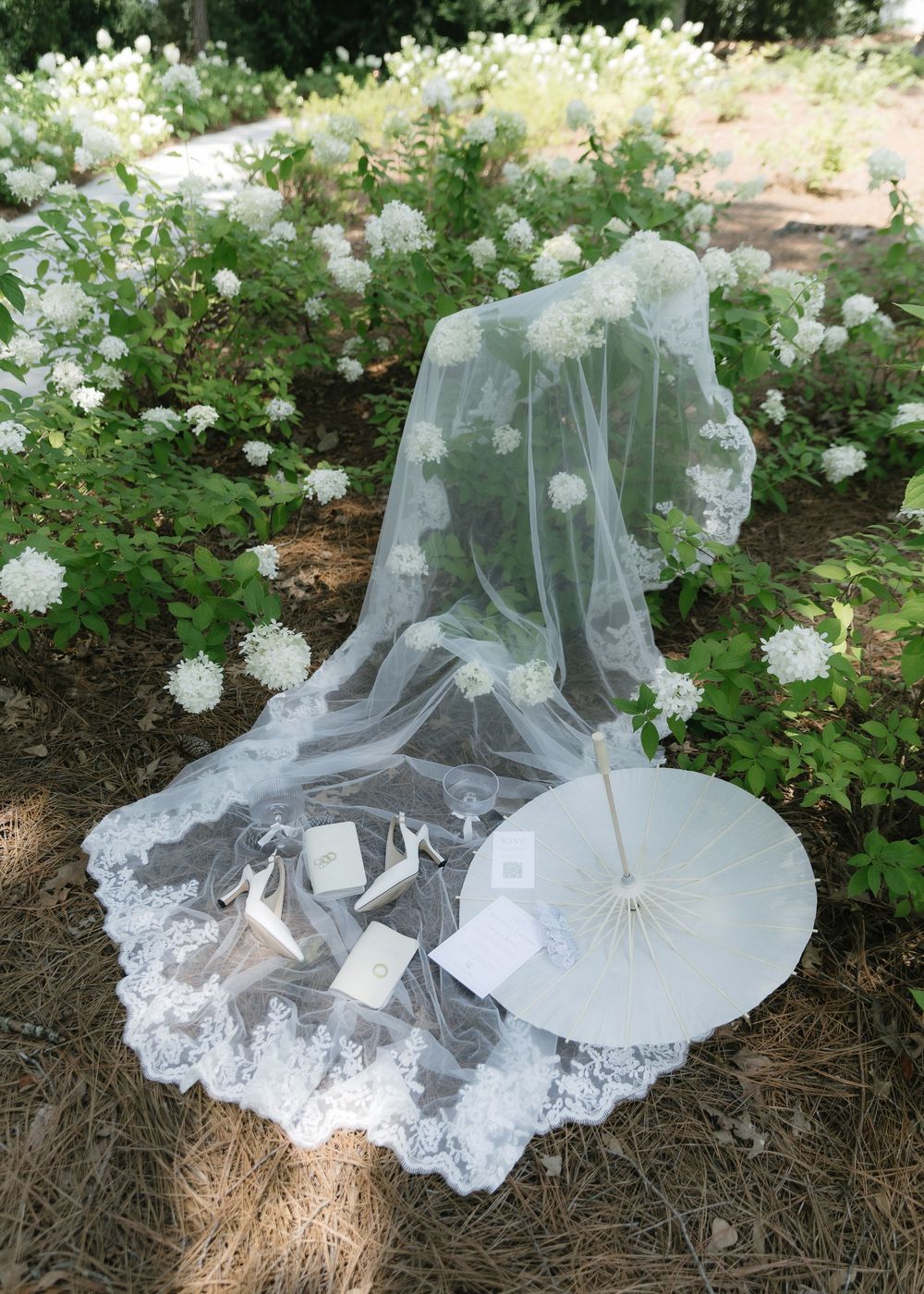 Bridal veil with lace trim and floral appliques laid on pine needles beside a white parasol and accessories in a garden.