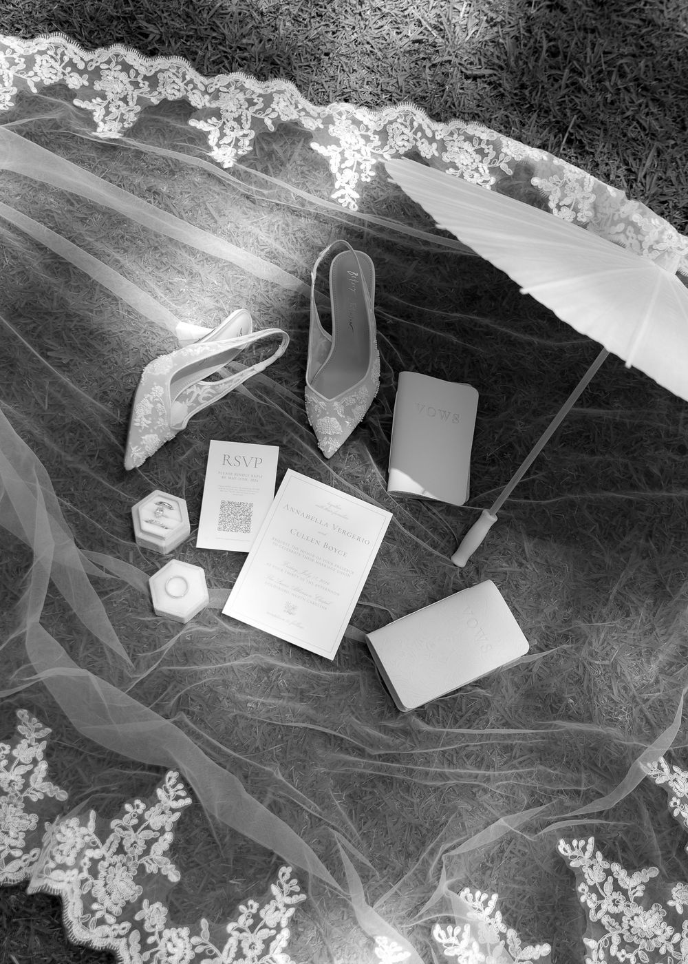 Black and white flat lay of bridal accessories including heels, veil, parasol, invitation suite, and ring box.