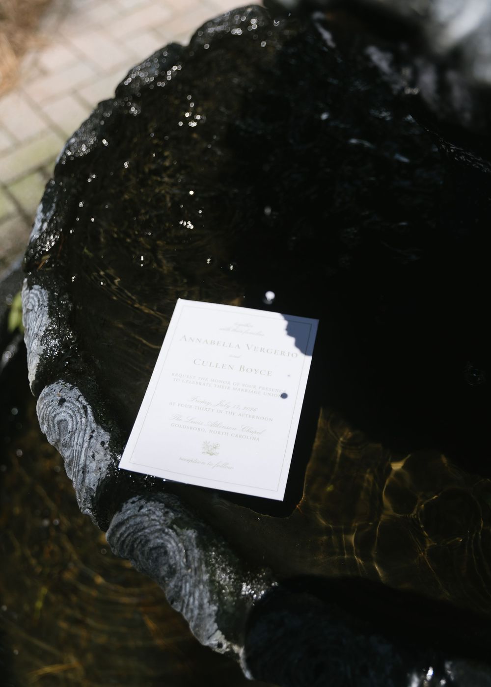 White wedding invitation card resting on a shiny black decorative stone surface outdoors.