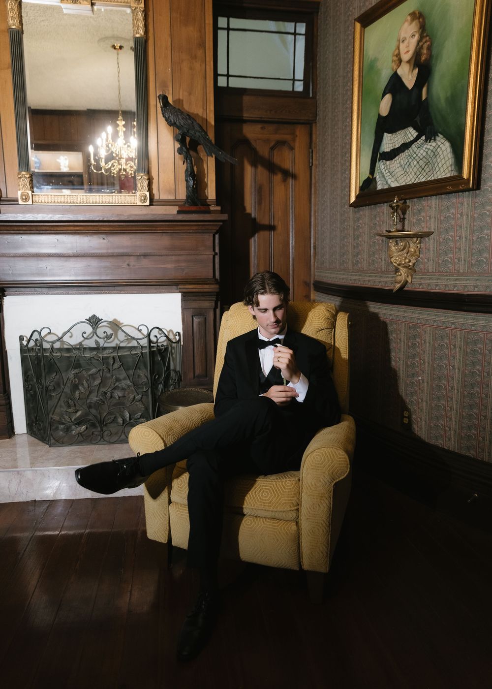 Man in tuxedo sitting in yellow armchair checking phone near fireplace in elegant wood-paneled room with portrait painting.