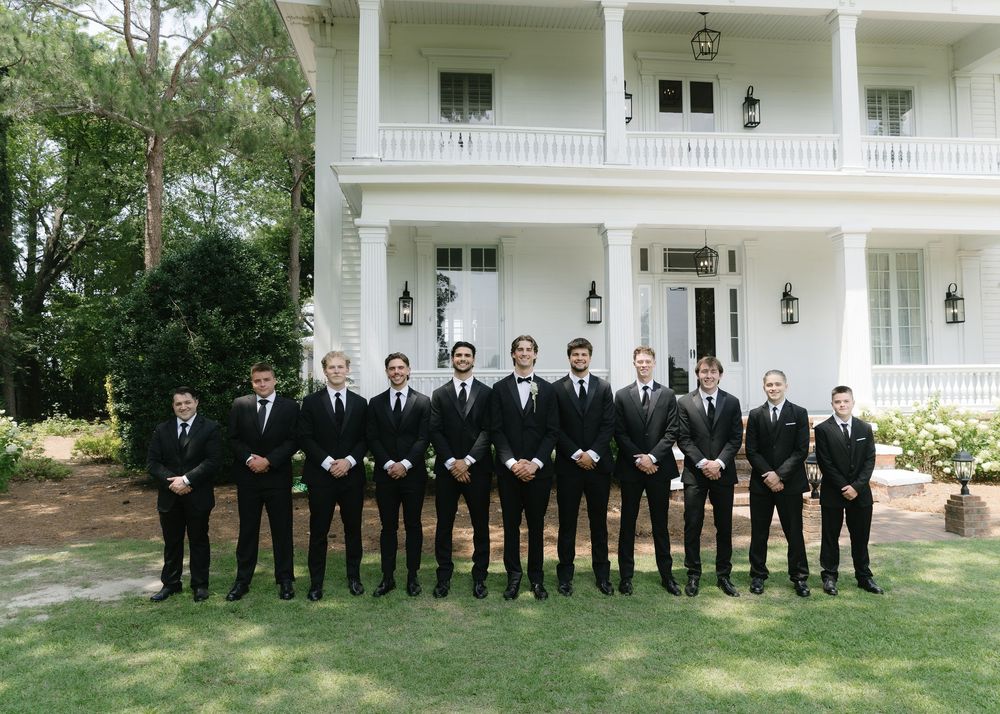 A group of groomsmen in black suits stand in a line on a lawn in front of a white two-story wedding venue.