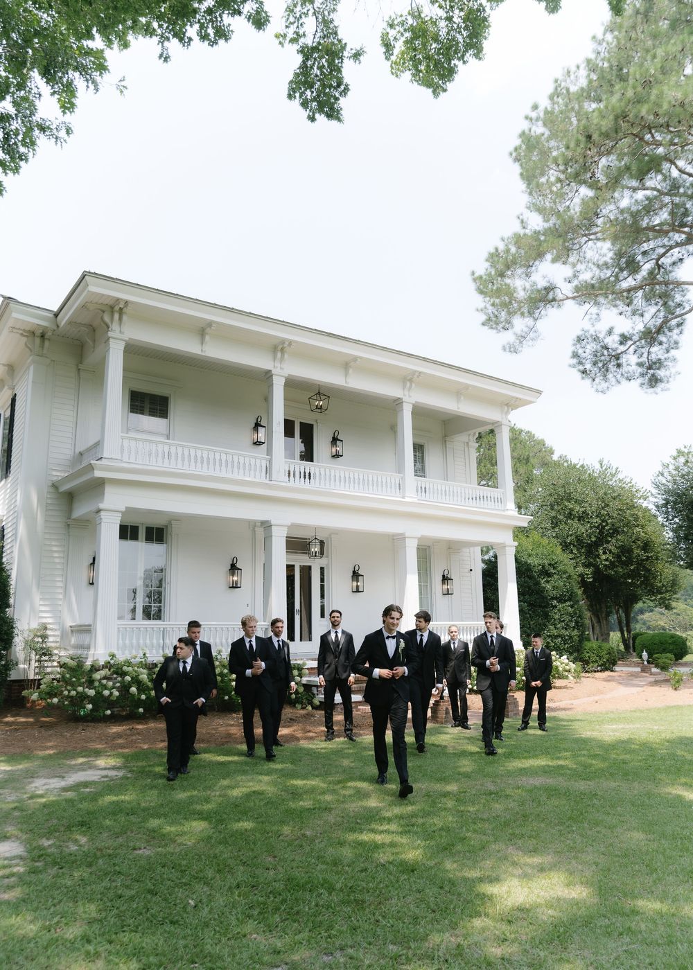 Groom and groomsmen in black suits walking across green lawn in front of elegant white two-story Southern mansion.