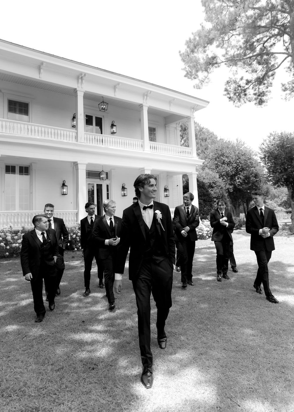 Groom leads groomsmen in tuxedos walking outdoors in front of elegant white Southern mansion in black and white photo.