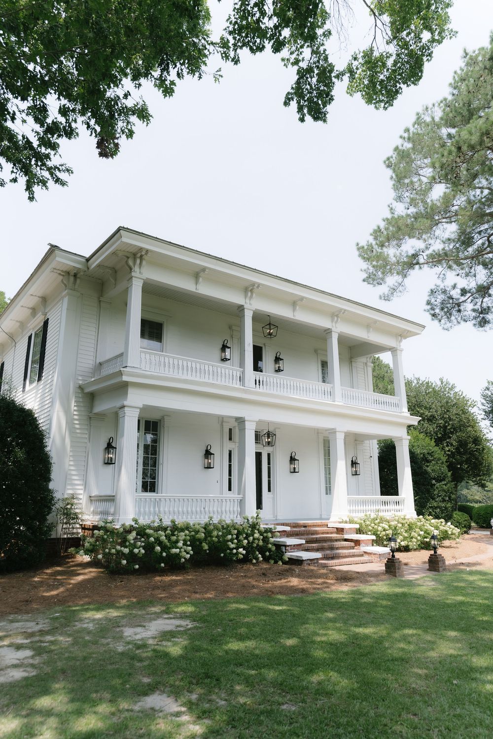 White two-story Southern colonial home with wrap-around porches, lantern lights, and lush green landscaping.
