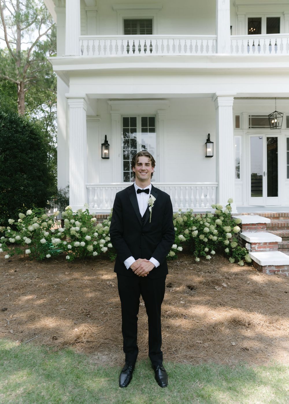 Groom in black tuxedo with boutonniere stands before elegant white Southern mansion with hydrangeas.