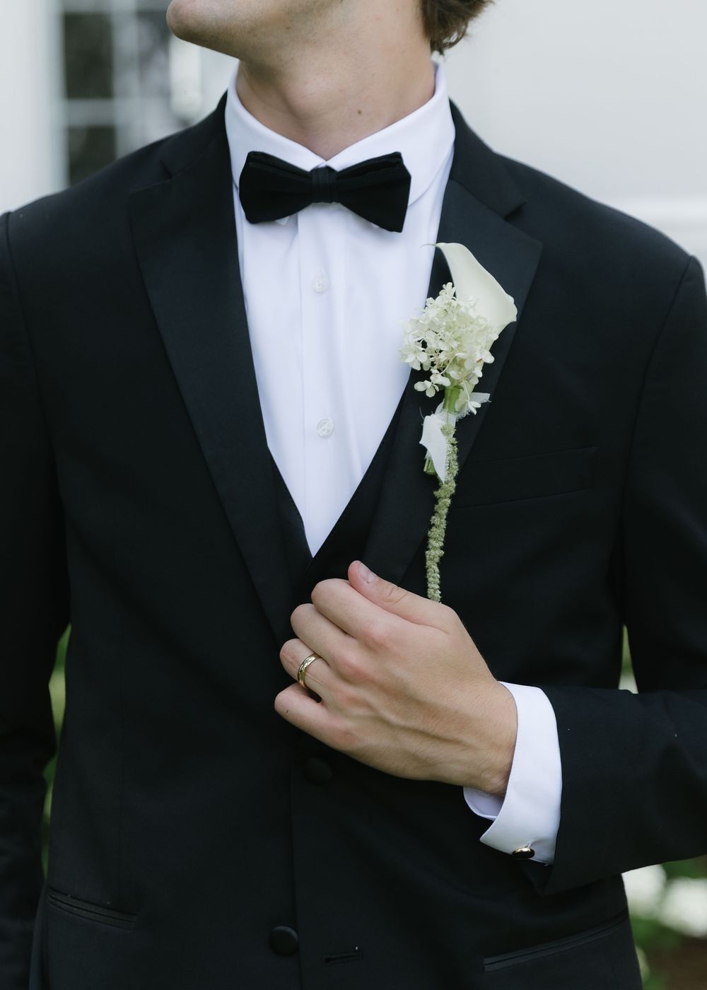 Groom in black tuxedo with bow tie and white floral boutonniere, wearing a gold wedding band.