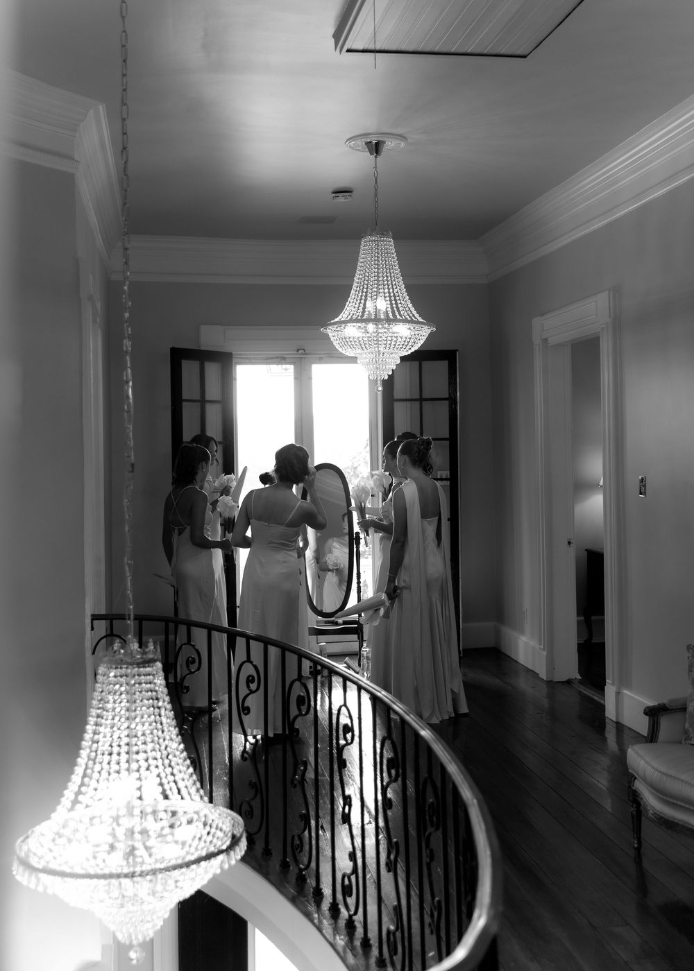 Black and white photo of bridesmaids helping bride in front of mirror in elegant hallway with crystal chandeliers.