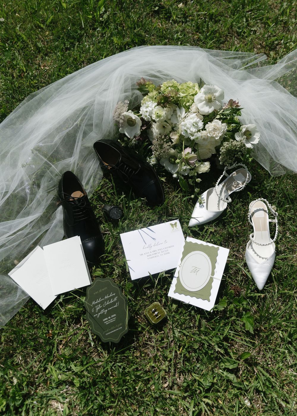 Wedding flat lay on grass featuring black oxford shoes, white heels, bridal veil, floral bouquet, and elegant green stationery.