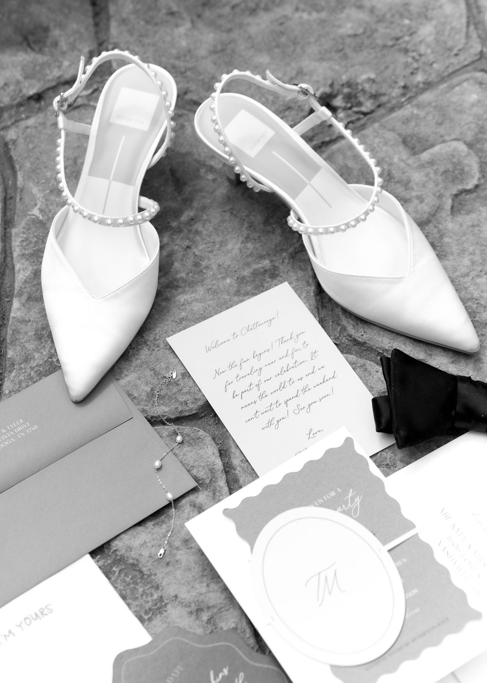 Black and white photo of pearl-adorned bridal heels beside elegant wedding stationery and a bow tie on stone.