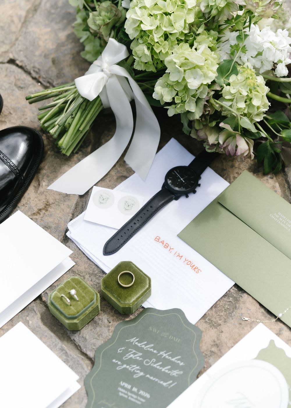 Wedding flat lay with hydrangea bouquet, green velvet ring box, black watch, invitation suite, and dress shoes on stone surface.