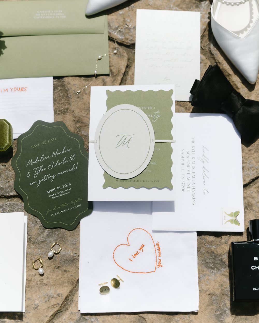 Elegant wedding invitation suite in sage green and white with monogram, heart doodle, and scattered jewelry on stone surface.