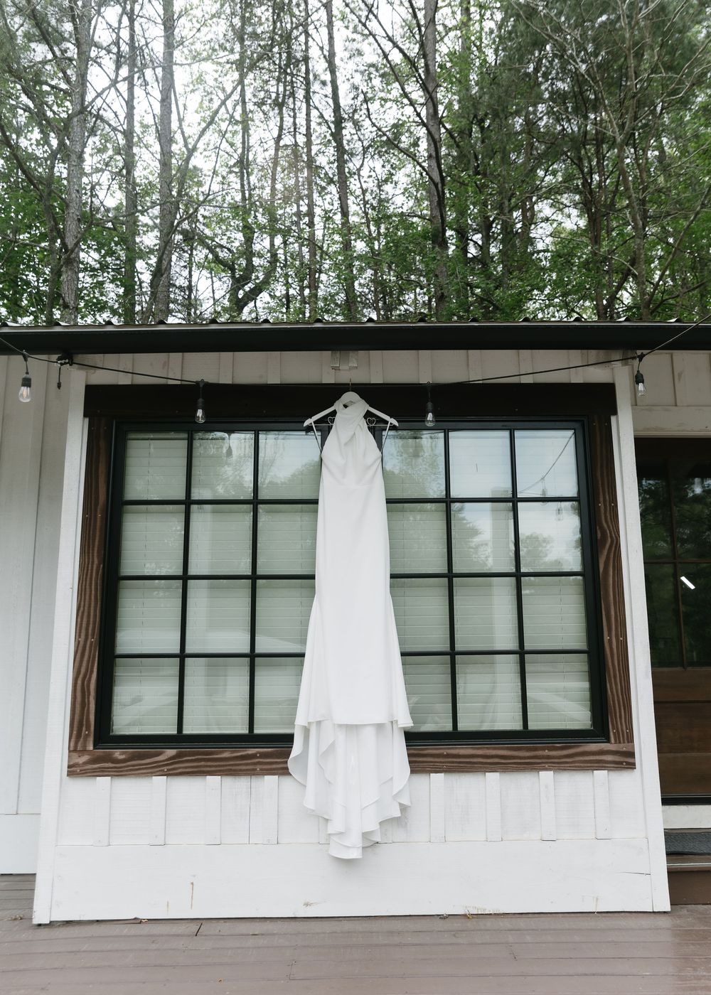 White wedding dress hanging on a hanger outside a white barn venue with large black-framed windows and string lights.