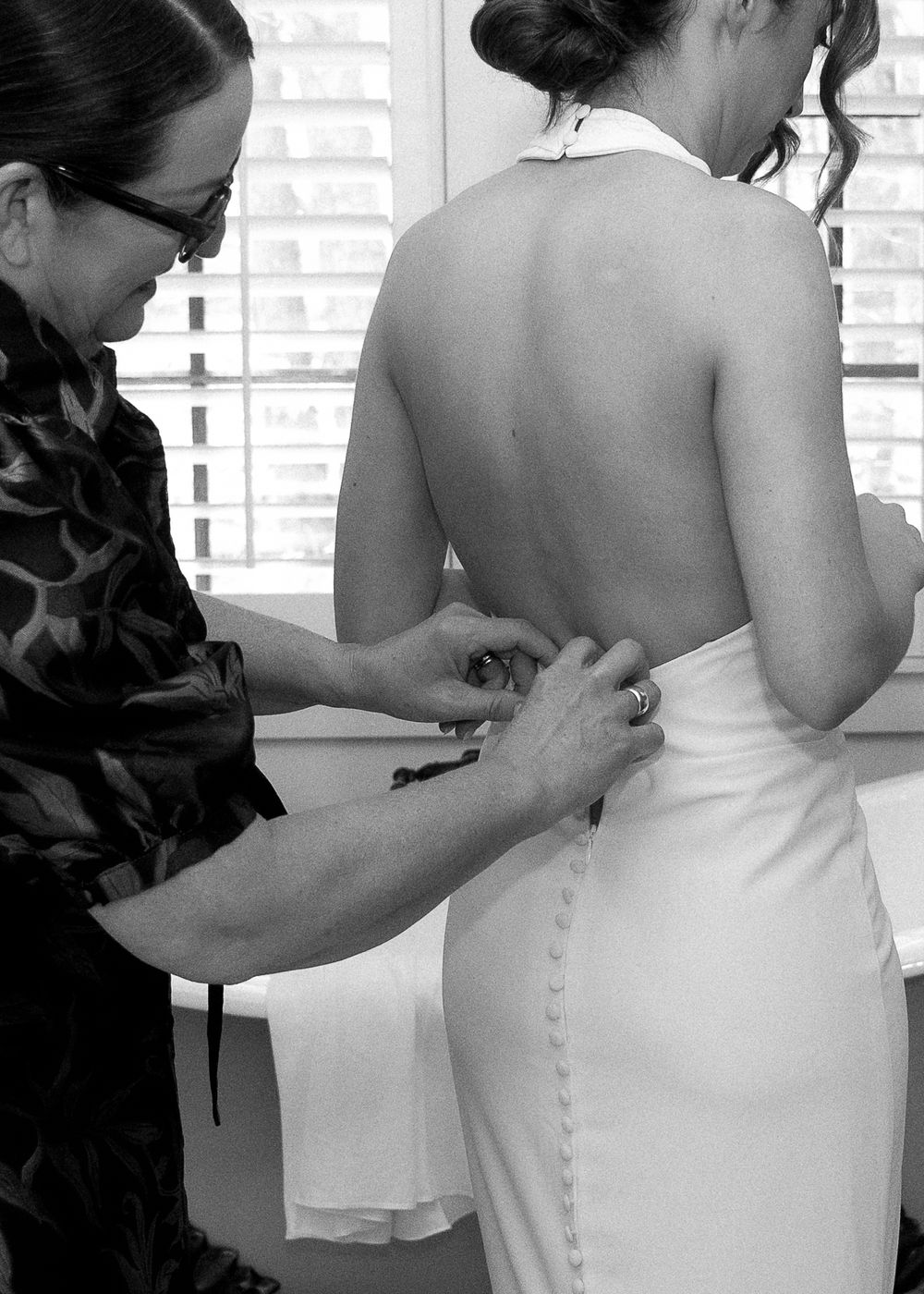 Black and white photo of a woman fastening buttons on a bride's backless halter wedding gown.