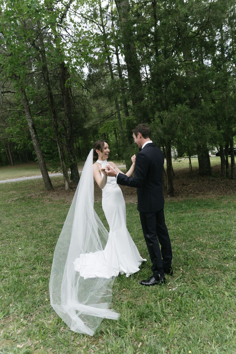 Bride in white gown and veil holds hands with groom in black suit outdoors on green grass surrounded by trees.