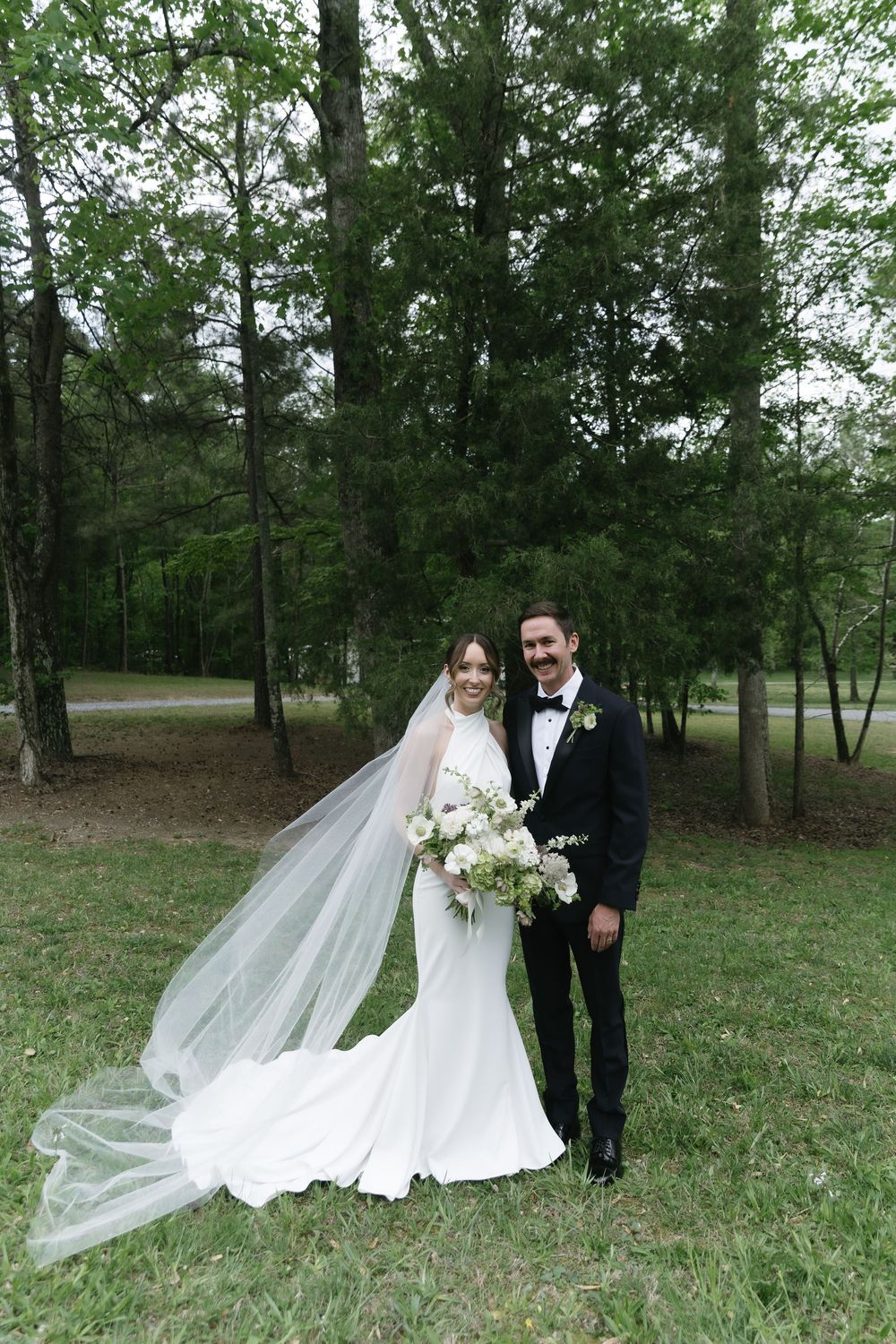 Bride in white gown with flowing veil holds bouquet beside groom in black tuxedo outdoors.