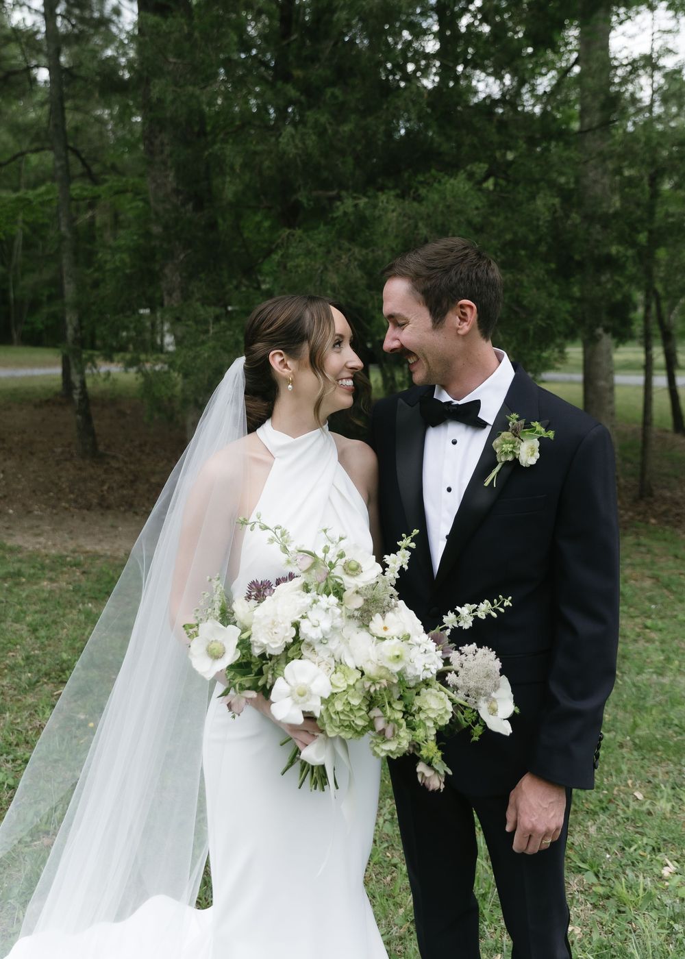 Bride and groom smiling at each other outdoors, she holds a white floral bouquet, he wears a black tuxedo with boutonniere.