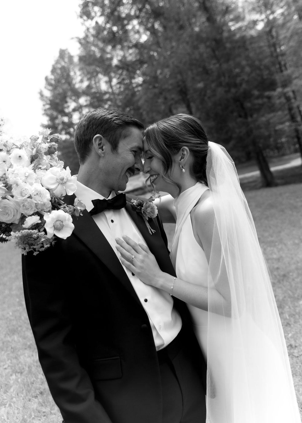 Black and white wedding photo of bride and groom touching foreheads, smiling, with floral bouquet outdoors.