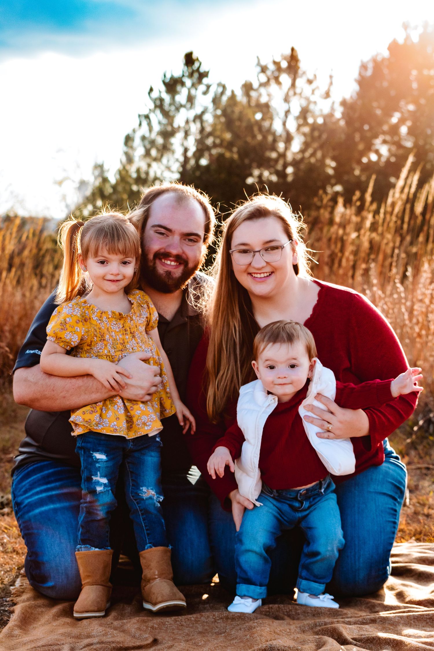 Young family of four poses outdoors in golden hour light, parents holding two toddlers in fall setting.