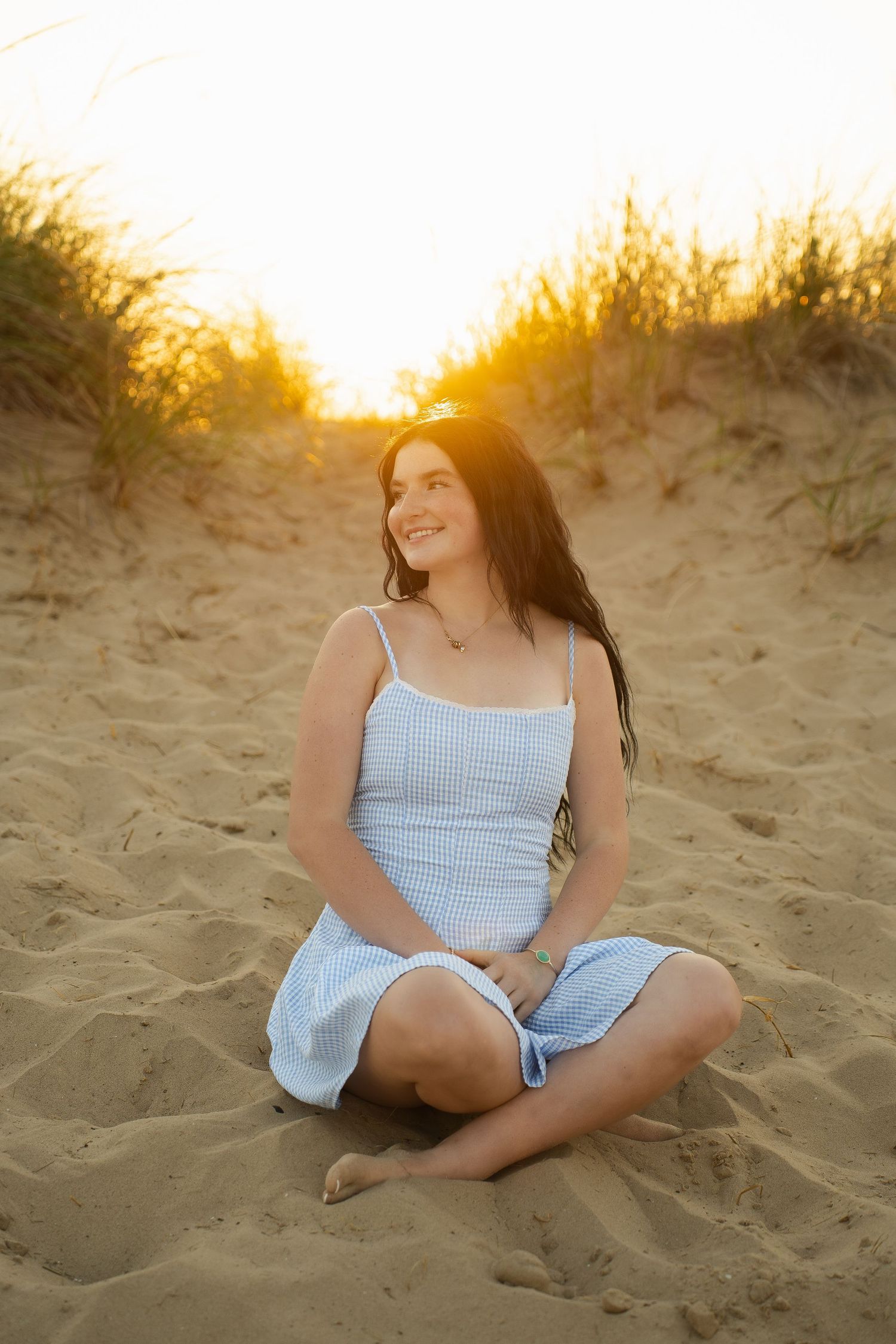 Young woman in blue sundress sitting cross-legged on sandy beach at golden sunset, smiling peacefully.