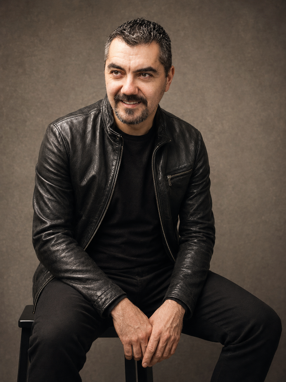 A confident middle-aged man in a black leather jacket sits on a stool, smiling against a neutral studio background.
