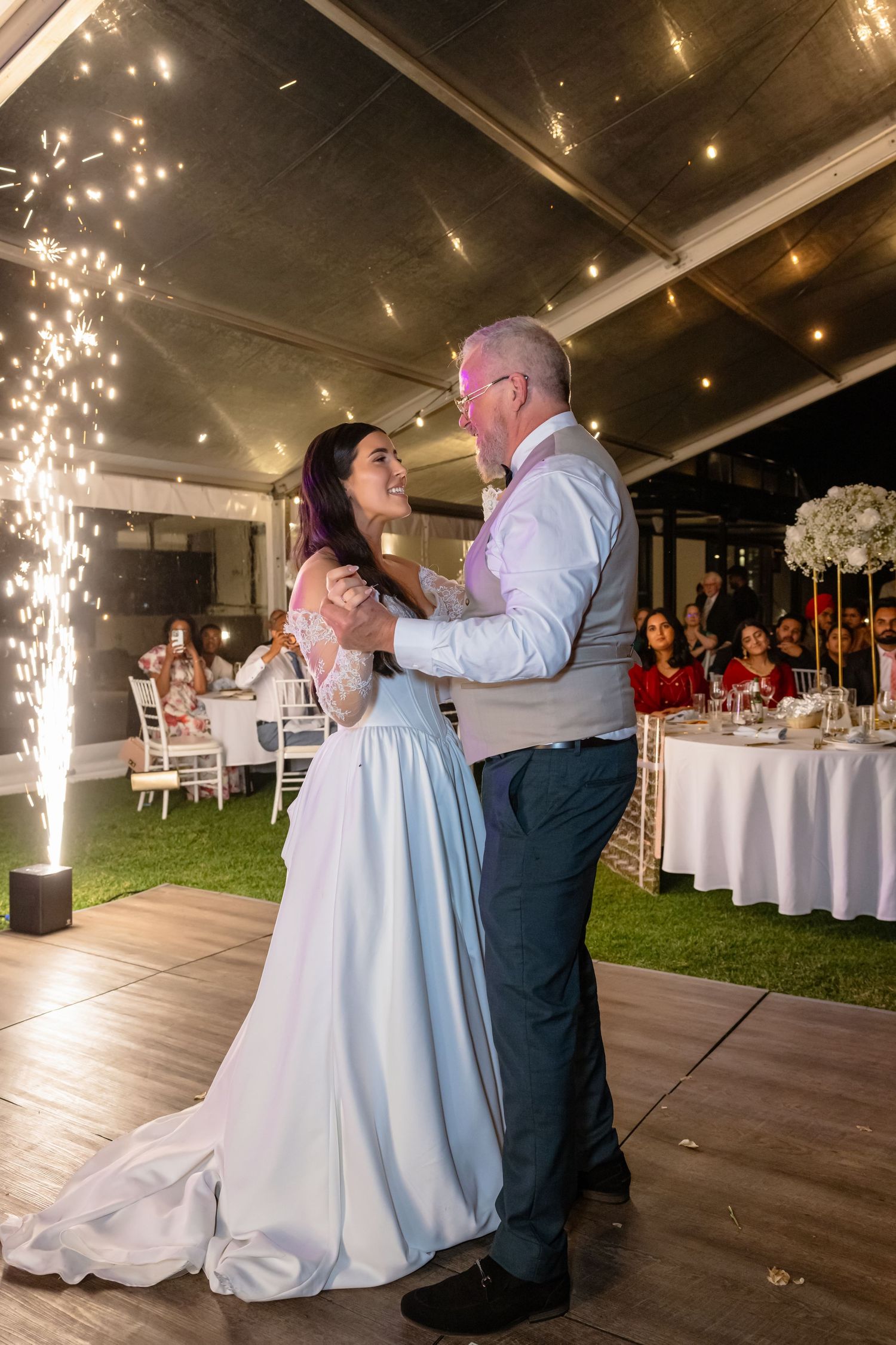 Bride and groom share first dance with sparklers at elegant outdoor wedding reception under string lights.