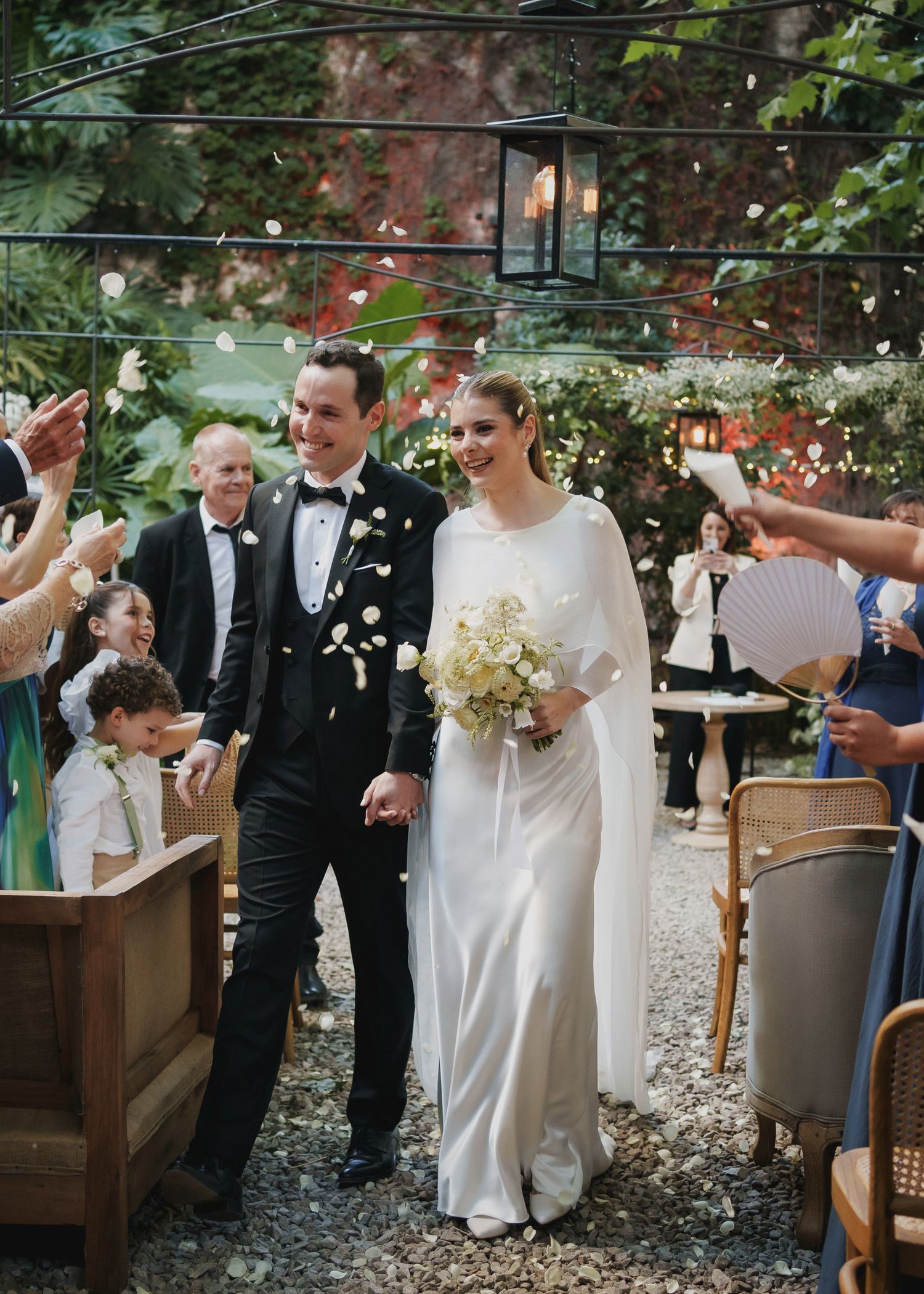 Bride and groom walking through confetti shower at lush garden wedding reception with string lights overhead.