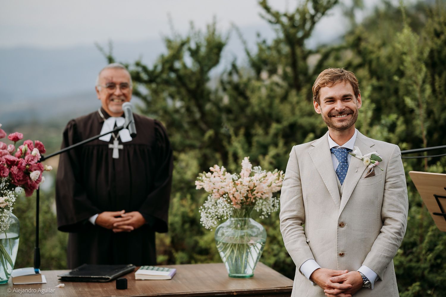 Smiling groom in beige suit stands at outdoor wedding altar with officiant and floral arrangements, mountains in background.