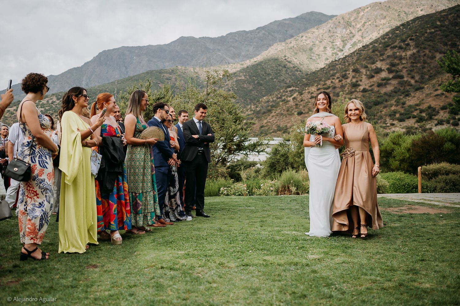 Bride walks down outdoor aisle with bridesmaid, guests applauding, mountain backdrop at scenic wedding.