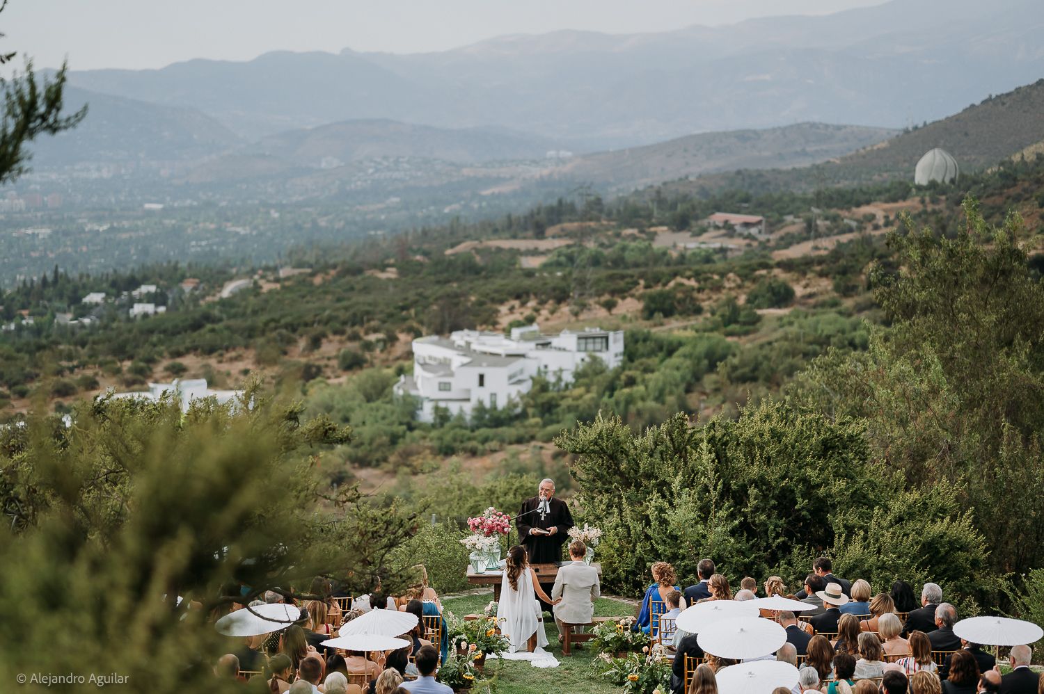 Outdoor wedding ceremony on a hillside with mountain views, guests seated under white umbrellas in a scenic landscape.