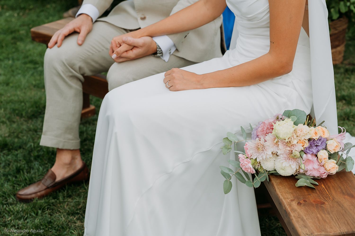 Bride and groom sit together on a bench, holding hands, with a pastel bouquet resting beside them.