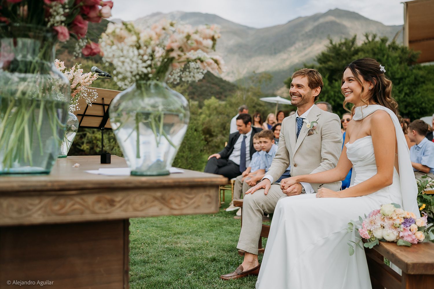Smiling bride and groom seated at outdoor wedding ceremony with mountain backdrop and floral decorations.