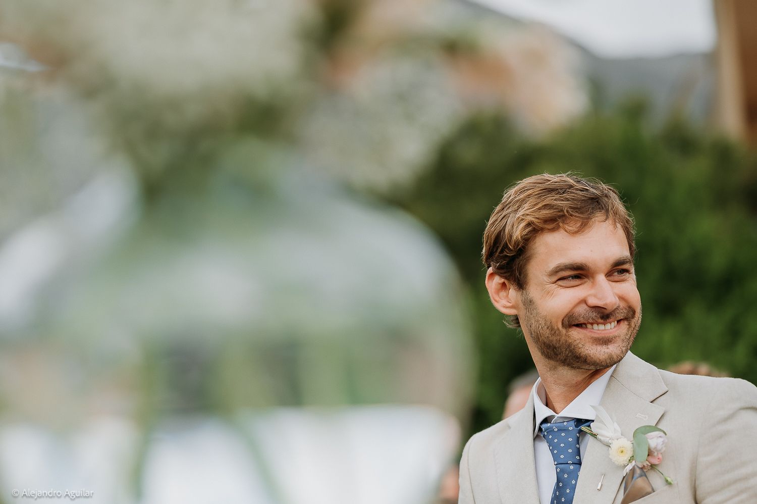 Smiling groom in beige suit with blue tie and boutonniere at outdoor wedding ceremony.