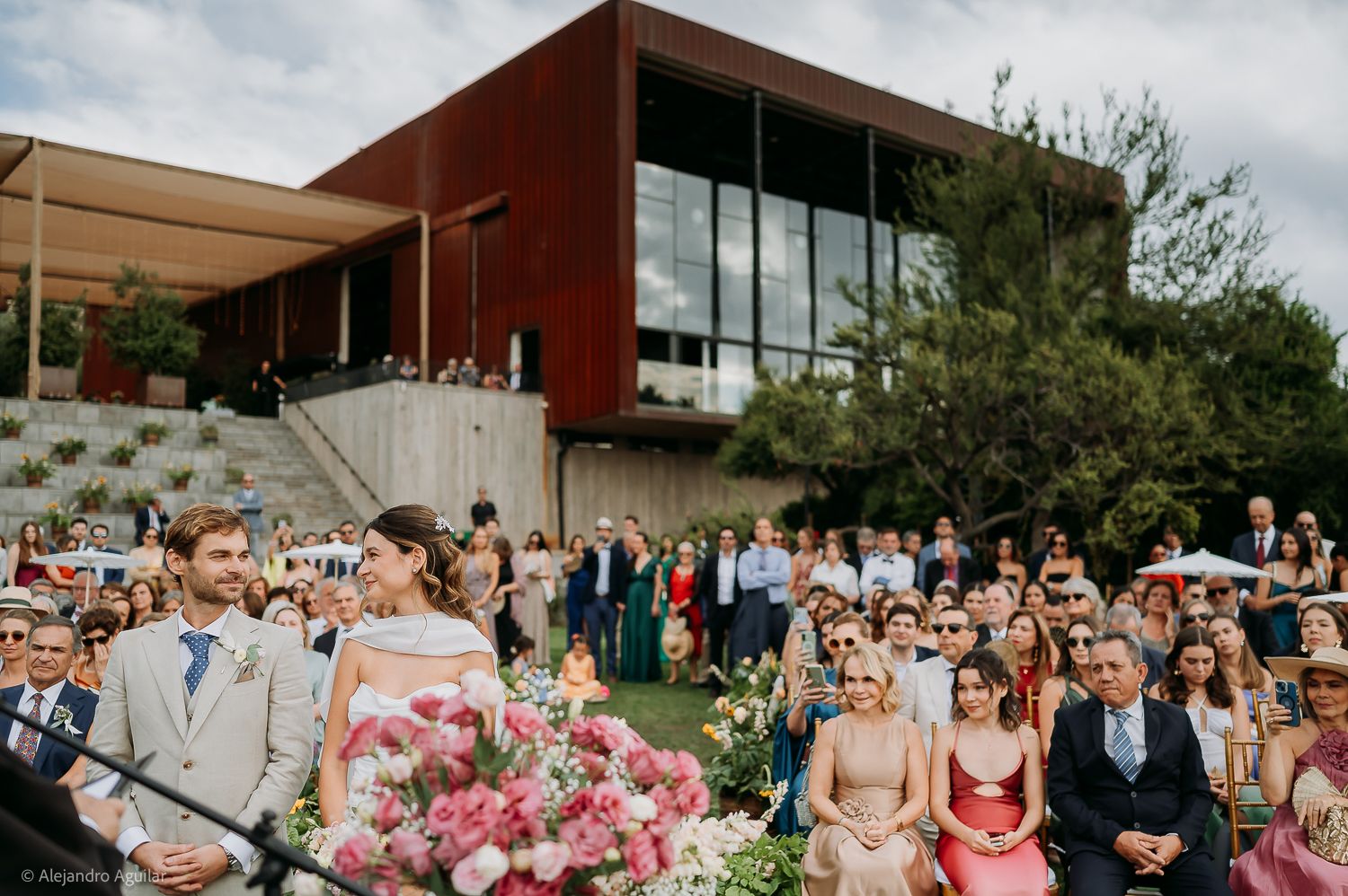 Outdoor wedding ceremony with bride and groom at altar, guests seated on lawn before modern building with red facade.