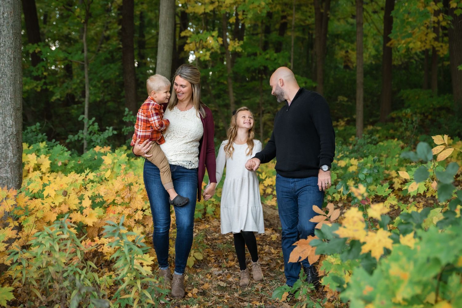 Family of four walking through colorful autumn forest, parents holding children among yellow and green fall foliage.