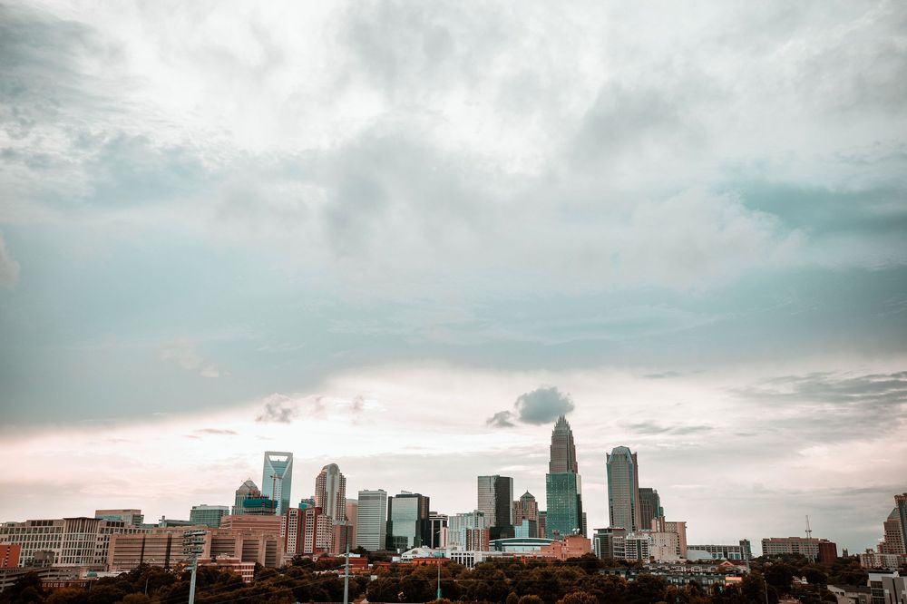 Charlotte NC city skyline with modern skyscrapers under dramatic cloudy sky surrounded by lush green trees.