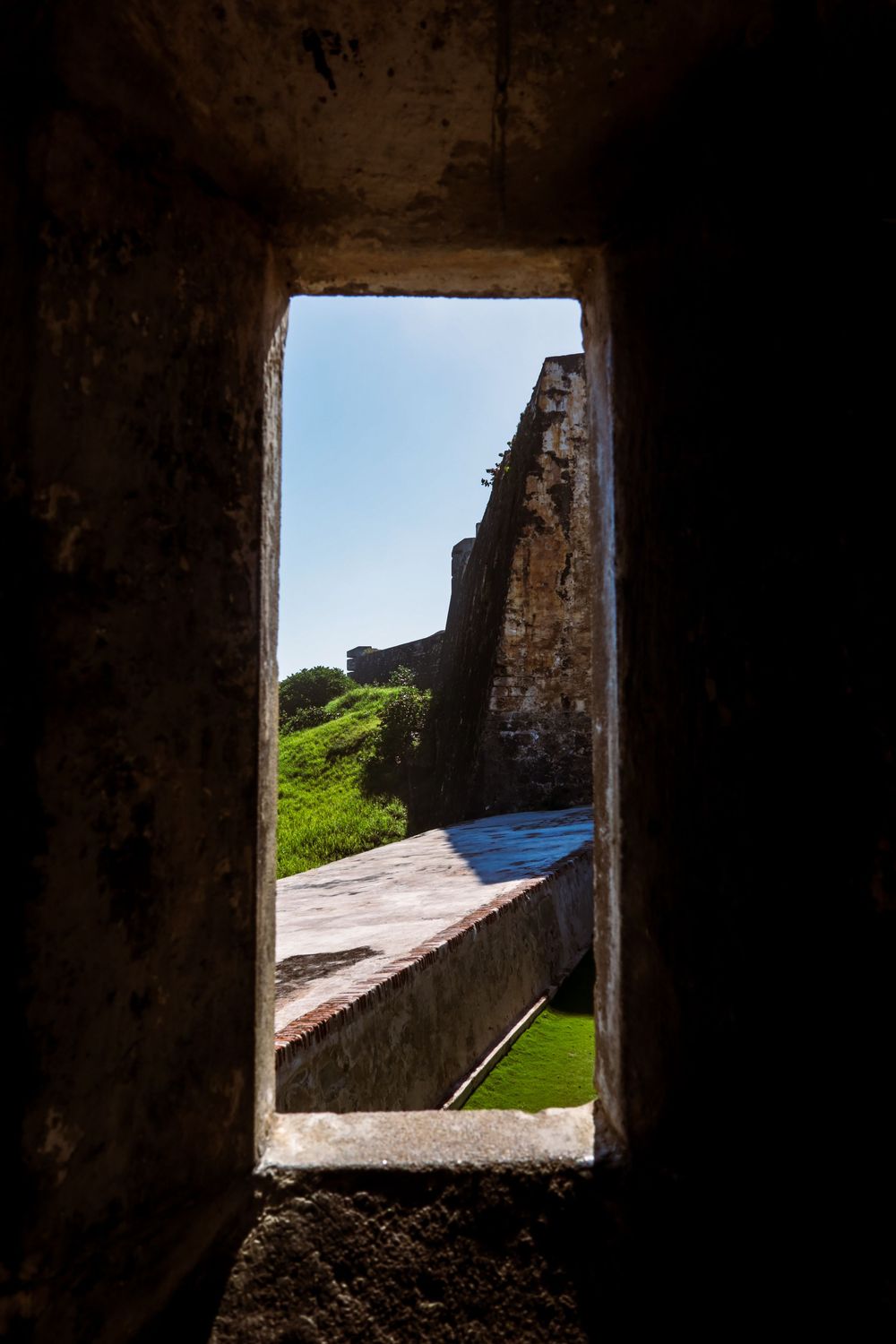 View through a narrow stone window of an ancient fort wall with green grass and blue sky beyond.
