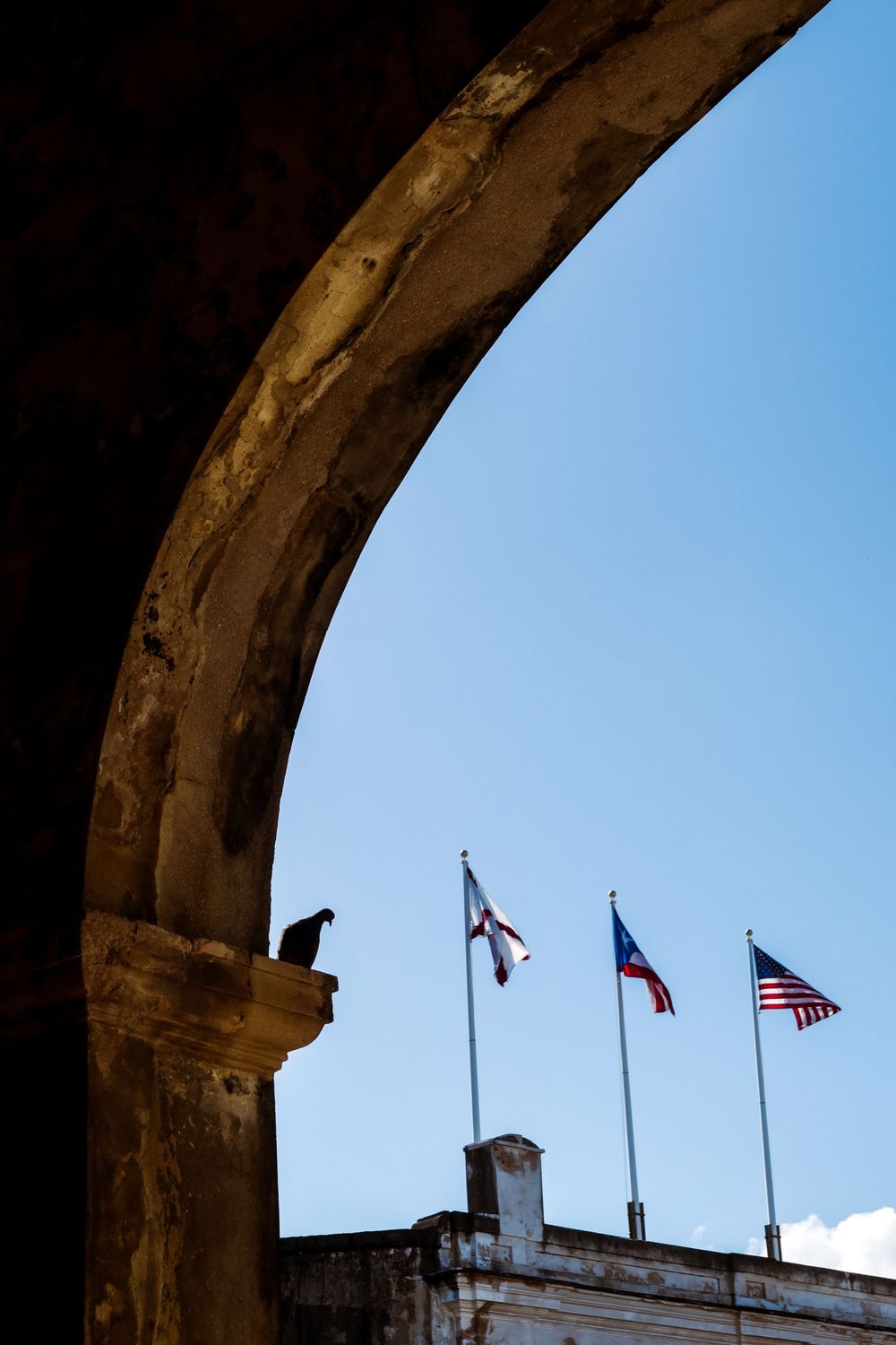 Stone arch frames three flags including US and Puerto Rico flags against blue sky, with a bird perched on the ledge.