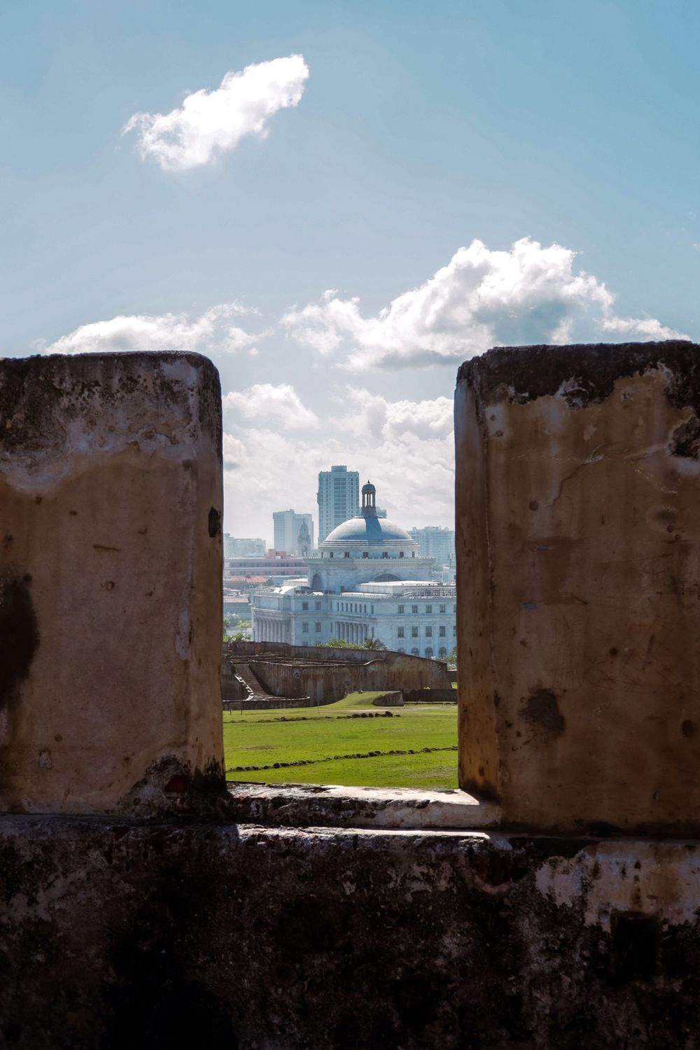 View through ancient fort battlements toward Puerto Rico Capitol building dome and modern city skyline under blue sky.