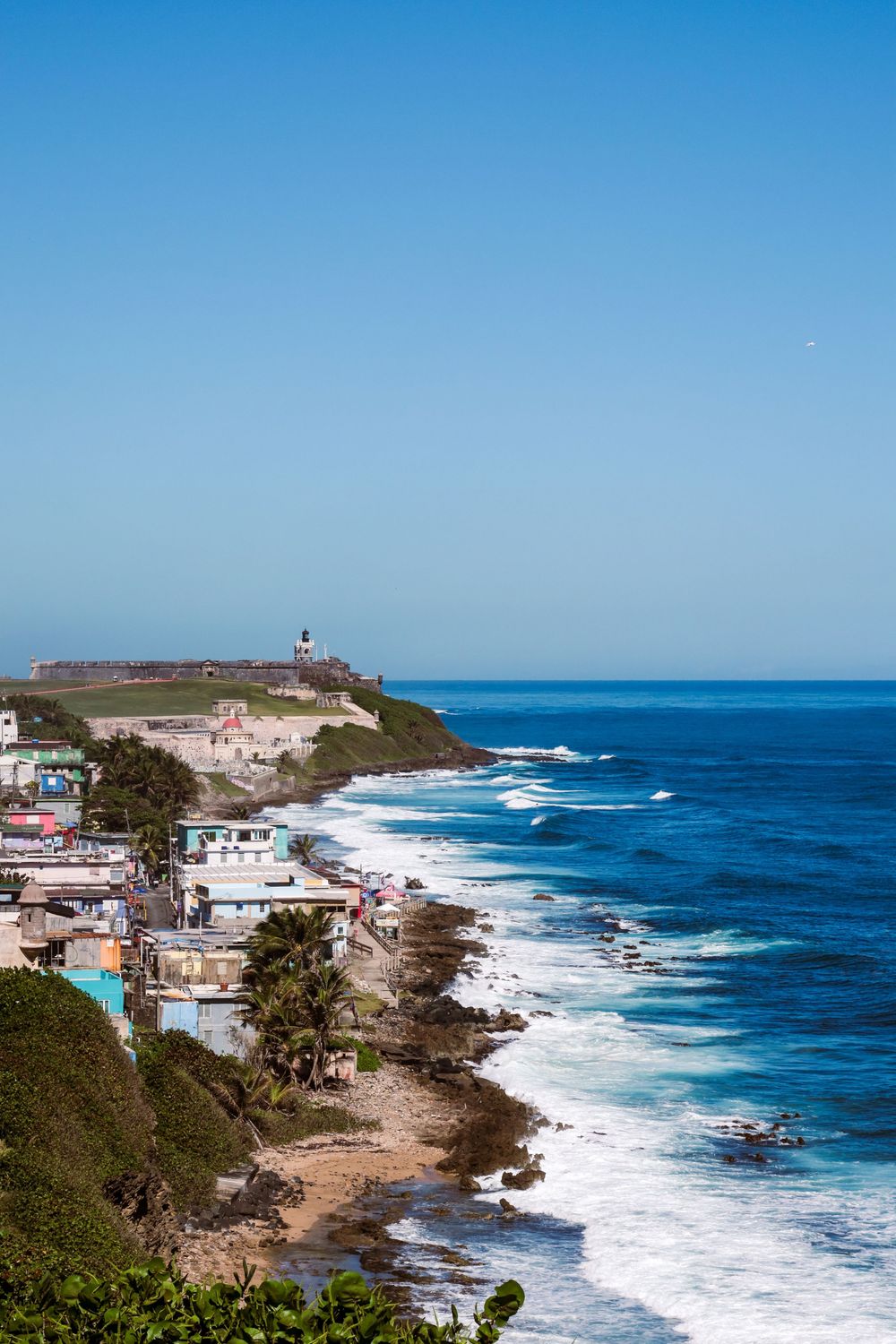 Aerial view of a rocky coastline with colorful buildings, crashing waves, and a lighthouse on a green cliff under a clear blue sky.