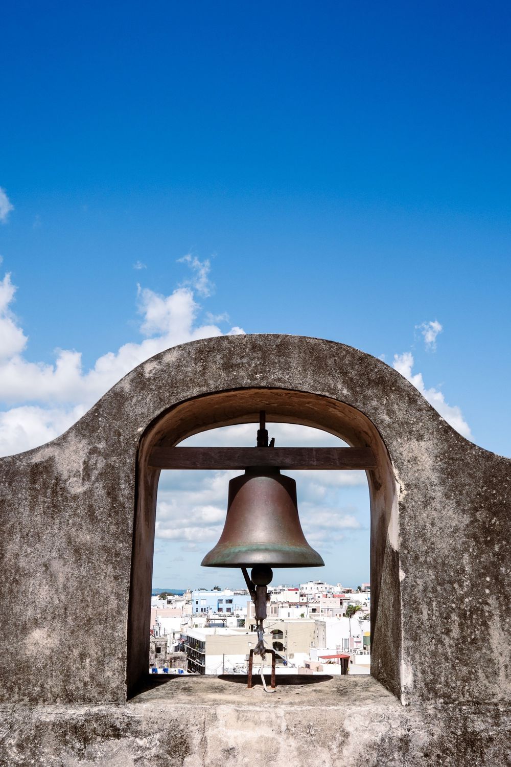 Bronze bell hanging in a stone arch atop a historic tower with a cityscape and blue sky in the background.
