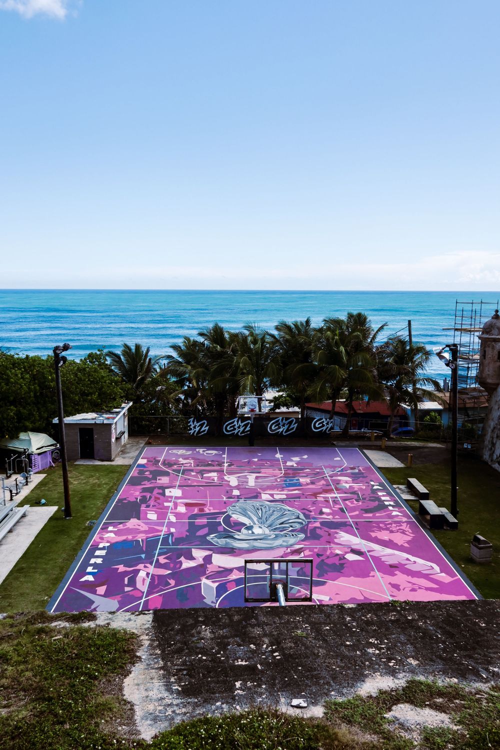 Aerial view of a vibrant pink and purple mural basketball court near a tropical beach with palm trees and blue ocean.
