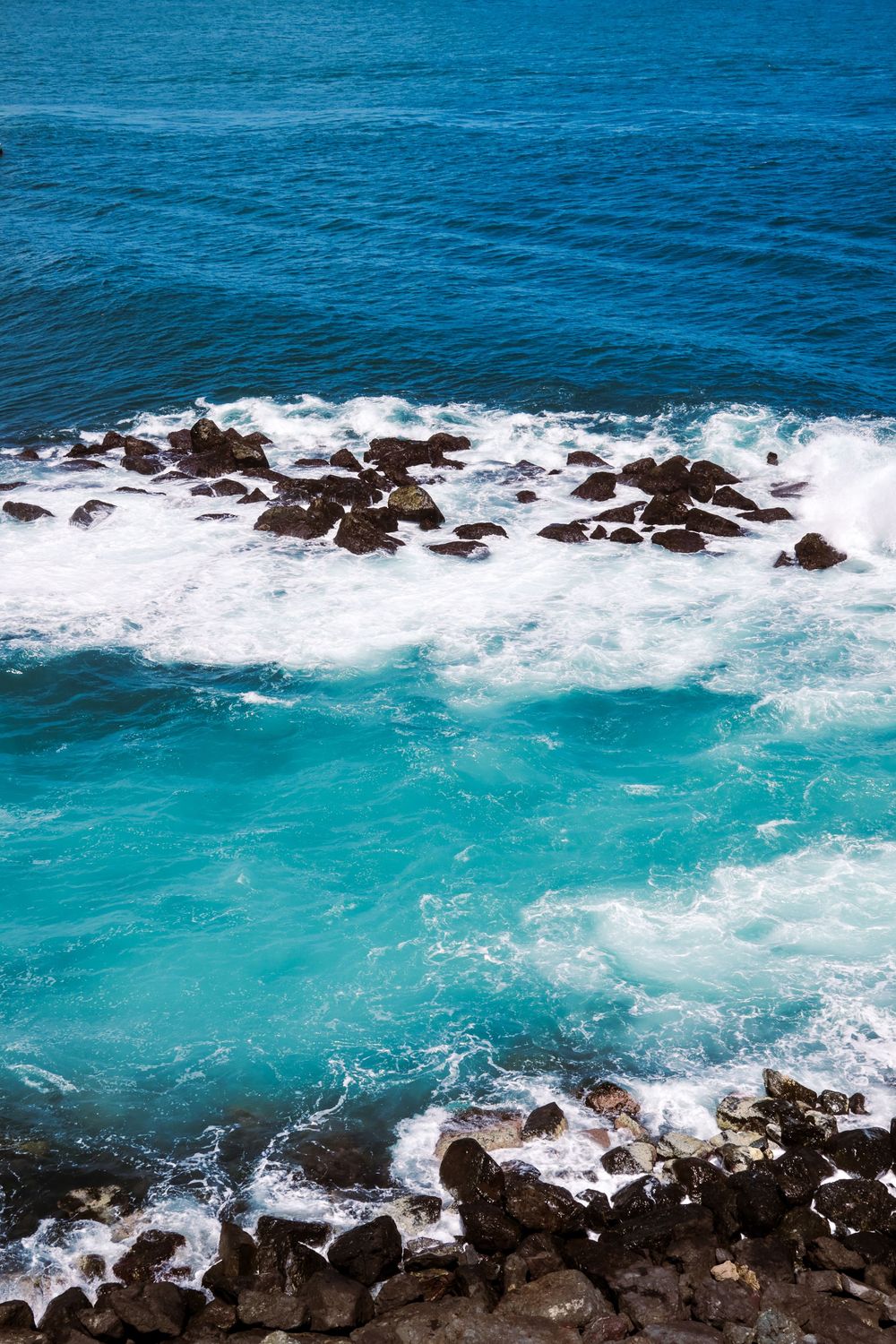 Aerial view of turquoise ocean waves crashing against dark volcanic rocks along a rugged coastline.