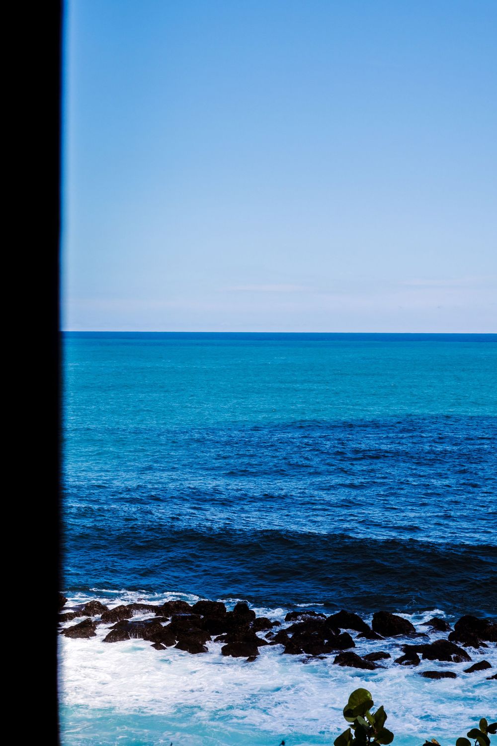 Blue ocean waves crashing on rocky shoreline viewed through window frame under clear sky.