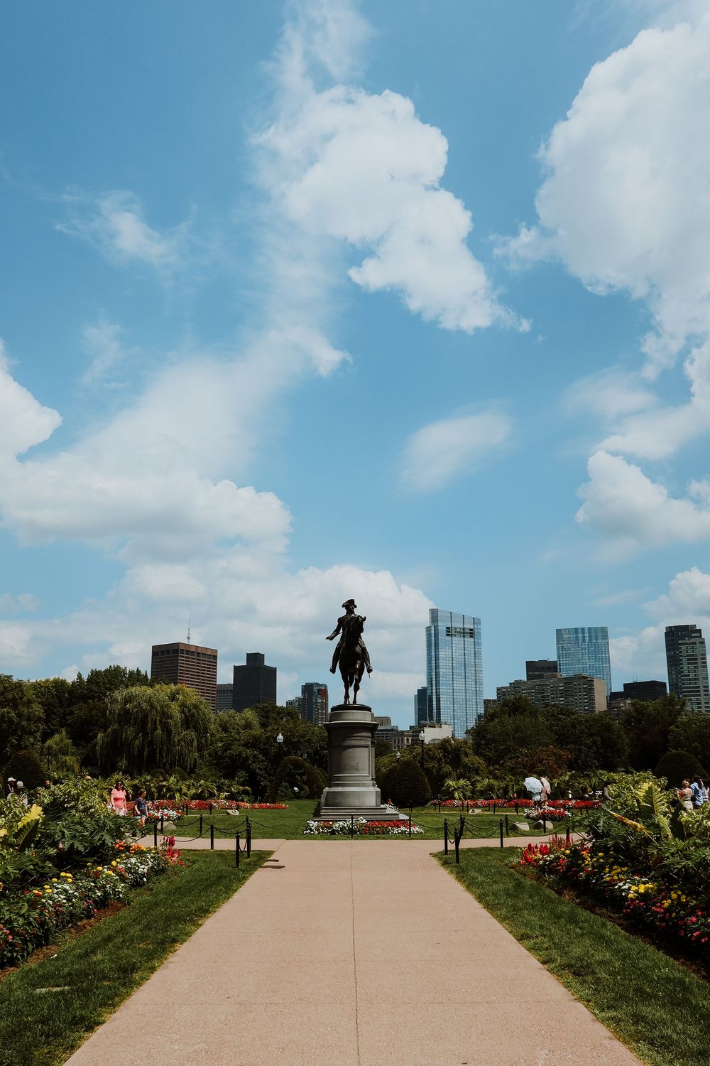 Equestrian statue in Boston Public Garden with colorful flower beds, lush trees, and city skyline under a partly cloudy blue sky.
