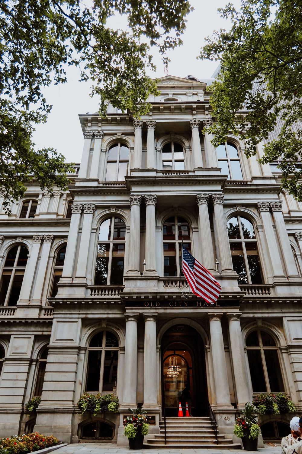 Historic neoclassical government building facade with American flag, arched windows, columns, and tree-lined entrance.