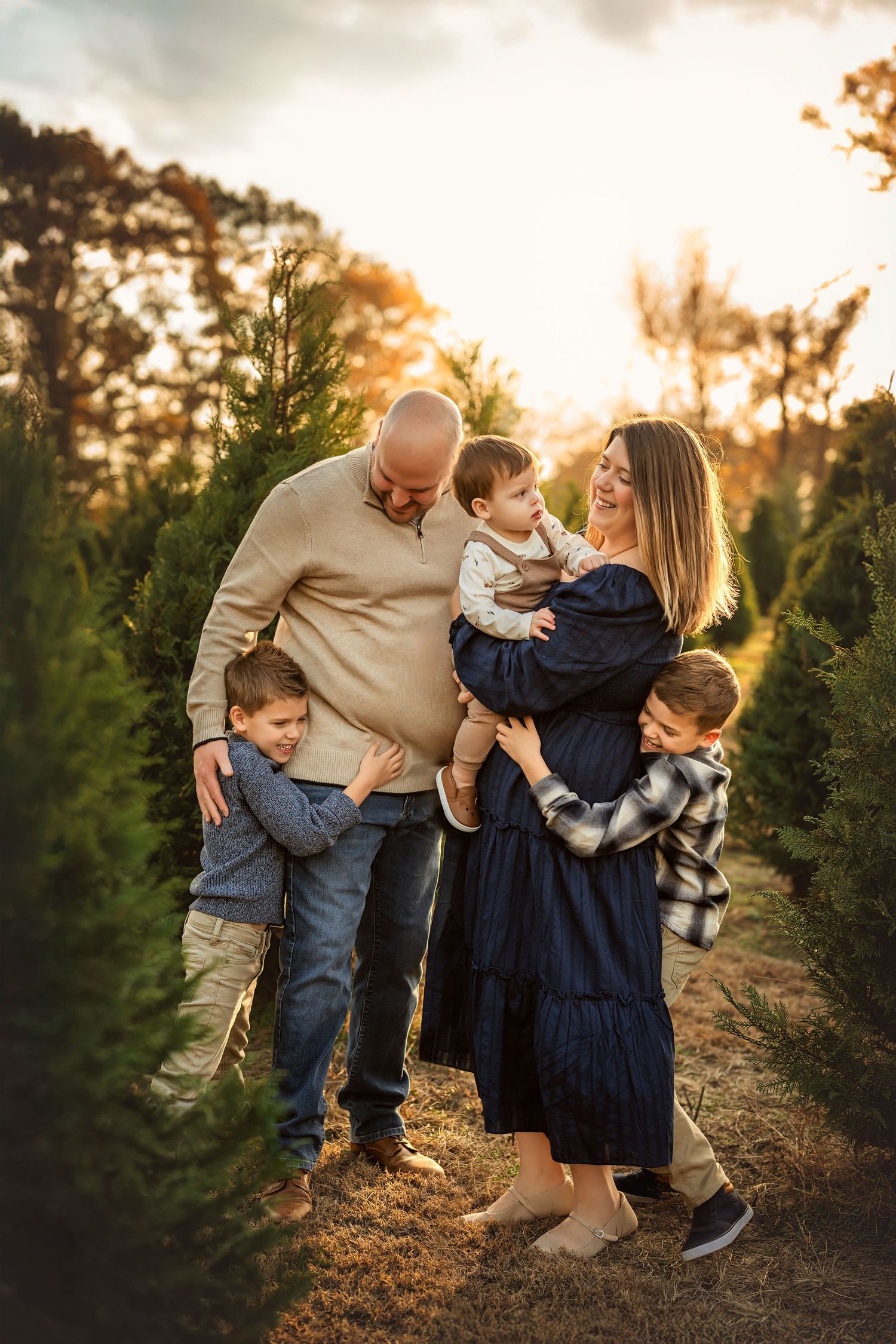 Family of five posing at sunset in a Christmas tree farm, parents holding children among evergreen trees.