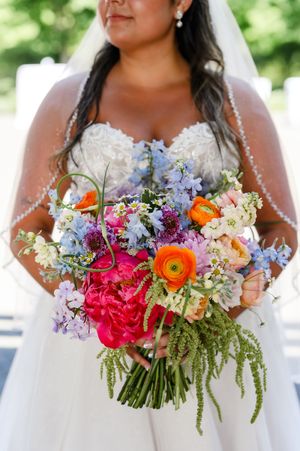 Bride in lace strapless gown holds vibrant bouquet with pink peonies, orange ranunculus, blue delphinium, and wildflowers.